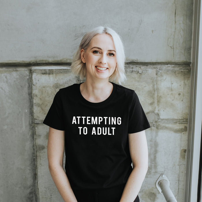 Woman with Blonde hair wearing black slogan tshirt which says Attempting to Adult